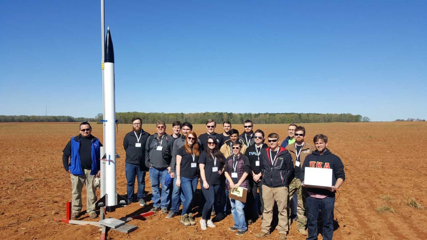 students stand next to a rocket primed to launch in a field