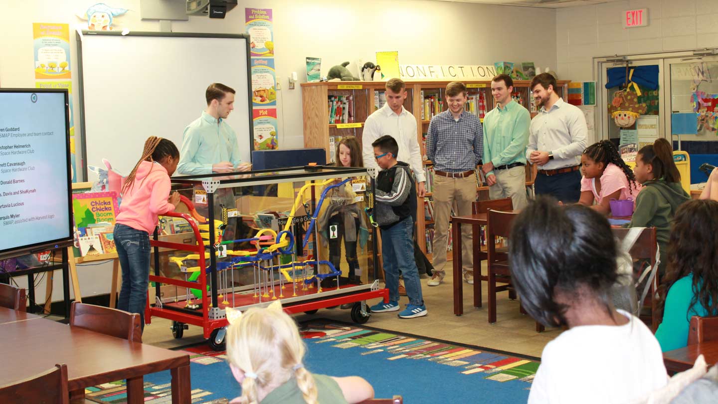 Students and children standing around the Mobile STEM Roller Coaster in an elementary classroom.