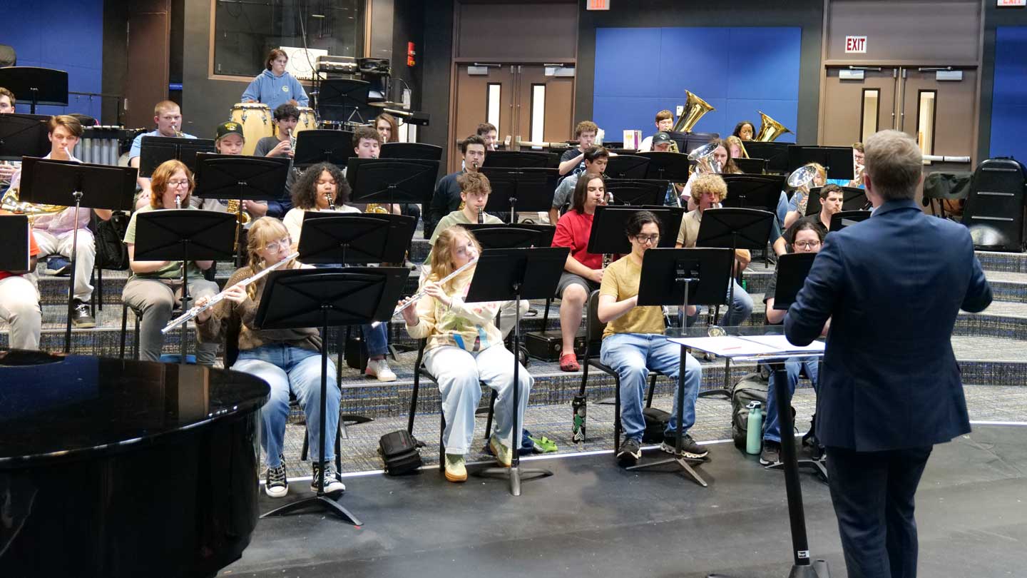 A music conductor leads a seated student band playing various instruments in a tiered rehearsal room.