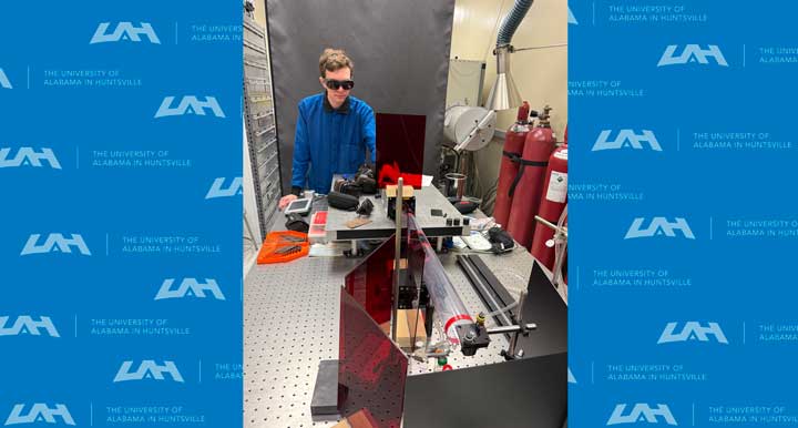 A person in a blue hoodie works at a lab table with scientific equipment, surrounded by gas canisters and safety gear.