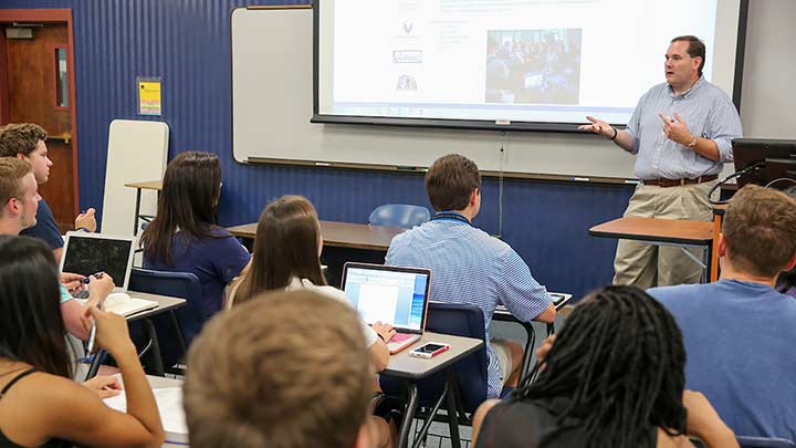 view of a classroom with teacher in front ?>