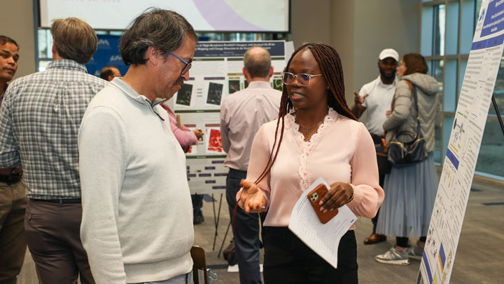 A UAH presenter engages in conversation while standing next to a research poster at an academic event. People network and discuss in the background.
