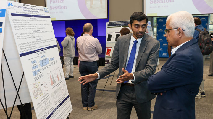 A UAH presenter explains a research poster to an attendee at a graduate poster session, surrounded by various research displays.
