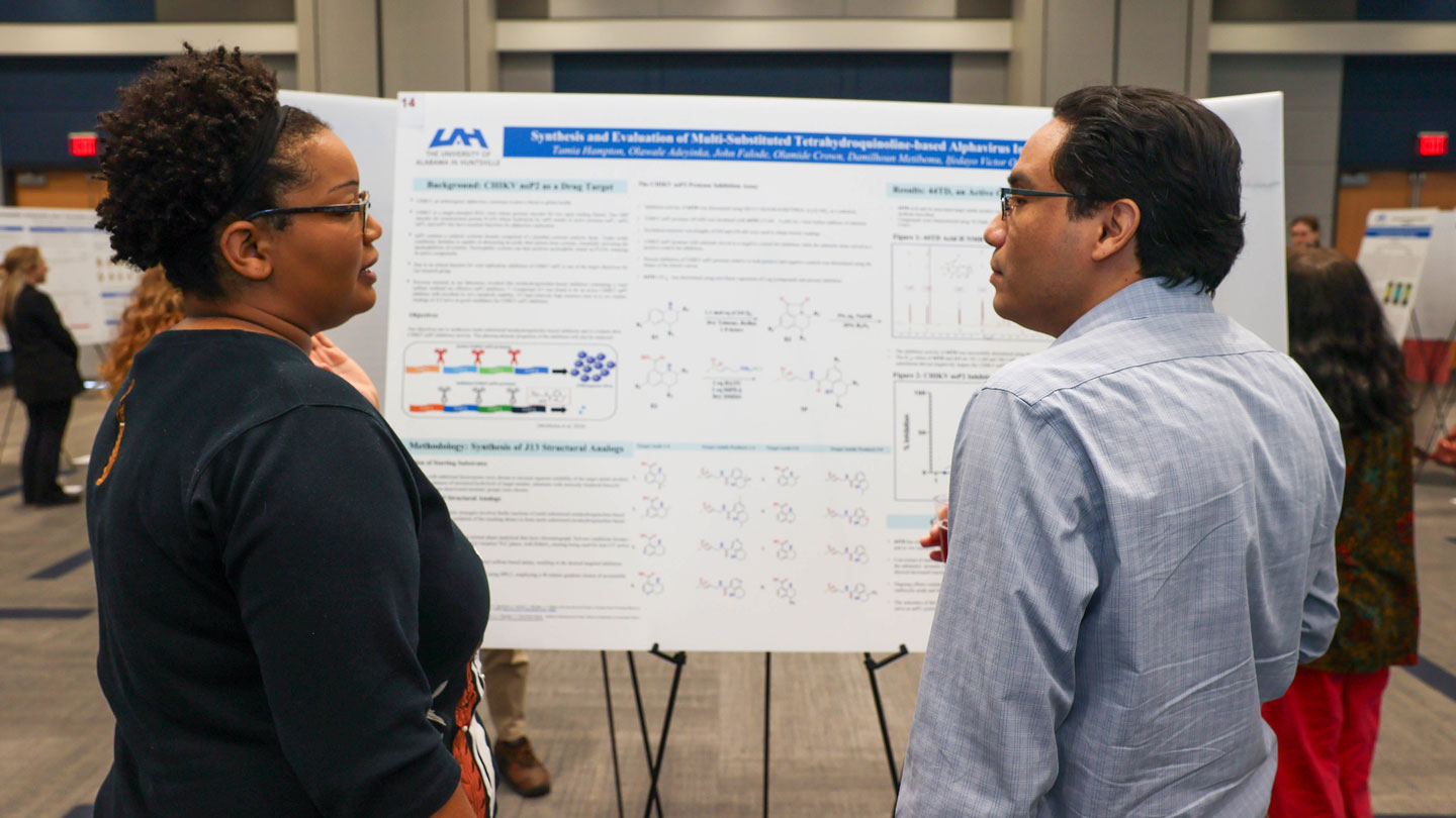 Two attendees engage in discussion in front of a scientific poster detailing molecular synthesis methods at a session.