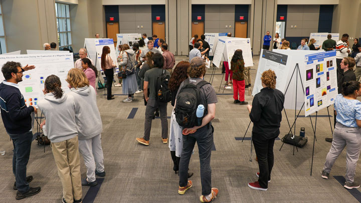 A busy conference room filled with attendees engaging with research posters on various topics, showcasing a collaborative academic event.