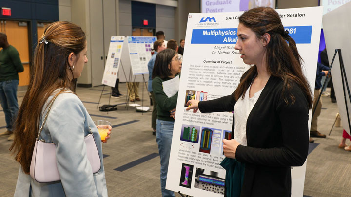 A graduate student presents a research poster titled Multiphyisics of Alkaline Batteries to an engaged person.