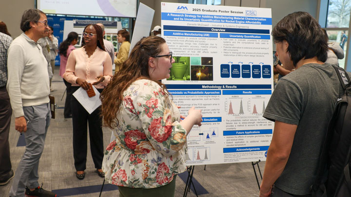 A graduate student presenting a research poster, surrounded by attendees engaging in discussion and viewing the poster.