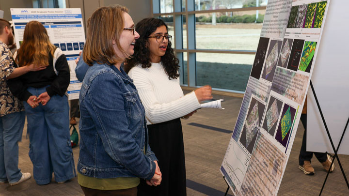A woman stands in front of a research poster, while others engage in discussions in a well-lit conference setting.