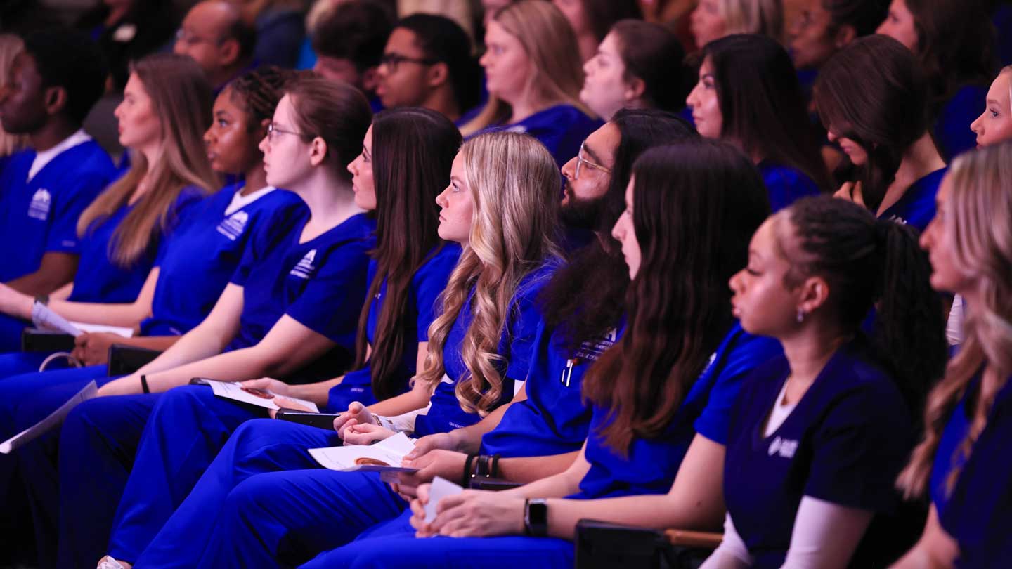 A group of UAH nursing students in blue uniforms sit attentively in rows in an auditorium.