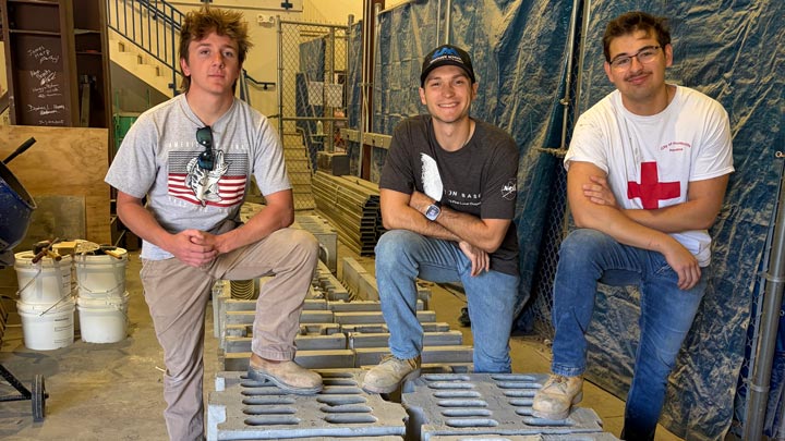 Three young men pose in a workshop, seated on stacked metal molds and surrounded by buckets, scaffolding, and blue tarps.