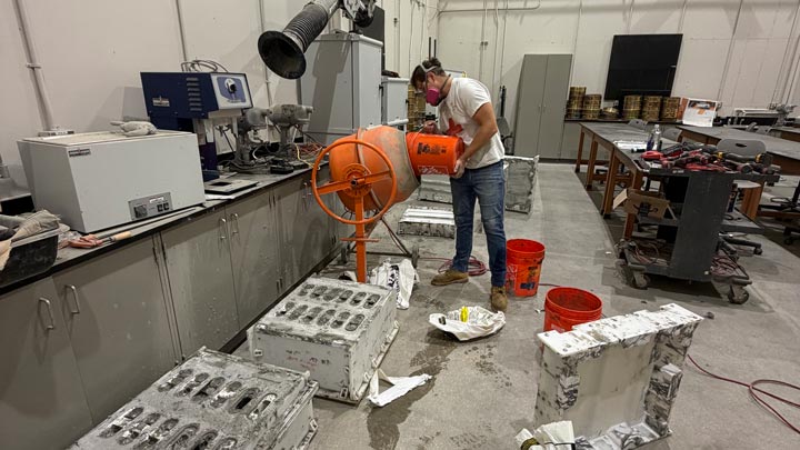 Worker operates an orange cement mixer in a laboratory filled with equipment, molds, tools, and buckets.