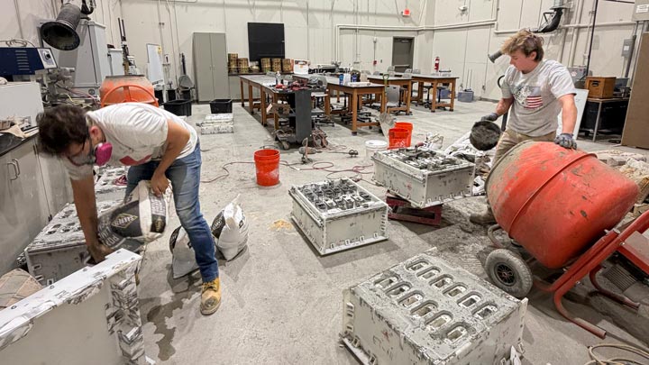 Two people work in an industrial workshop, handling equipment and materials among machinery, crates, and red barrels.