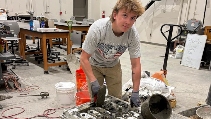 Young man in a workshop uses tools to work on a large metal engine component, surrounded by buckets, equipment, and worktables.