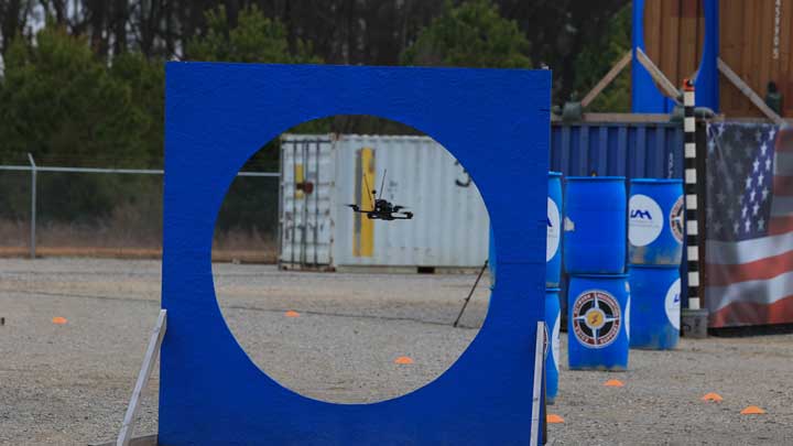 A drone flies through a large blue circular frame, set in an outdoor area with containers and barriers in the background.