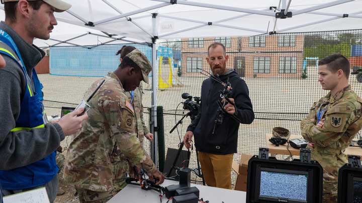 A military personnel inspects equipment at a tech demonstration under a tent, while others observe and take notes.