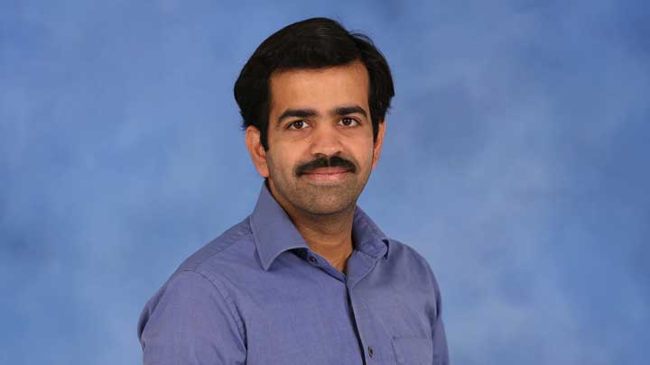 Studio portrait of Dr. Athiray Panchapakesan wearing a blue button-down shirt, standing against a blue backdrop and looking toward the camera with a neutral expression.