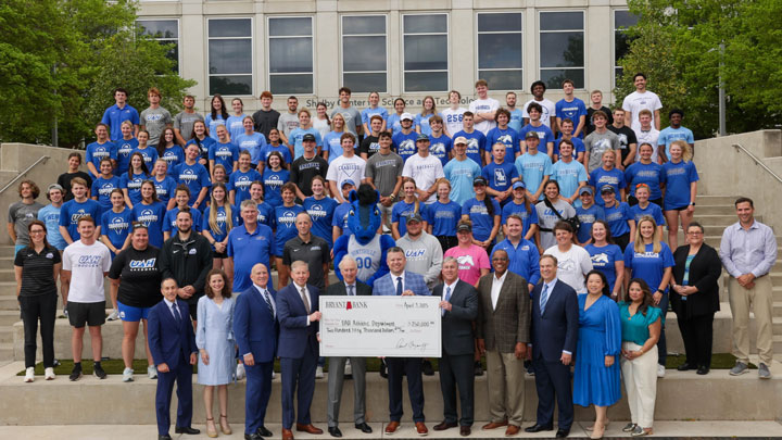 A large group of athletes and officials gather outdoors, holding a large check for $250,000, representing UAH's athletic department.