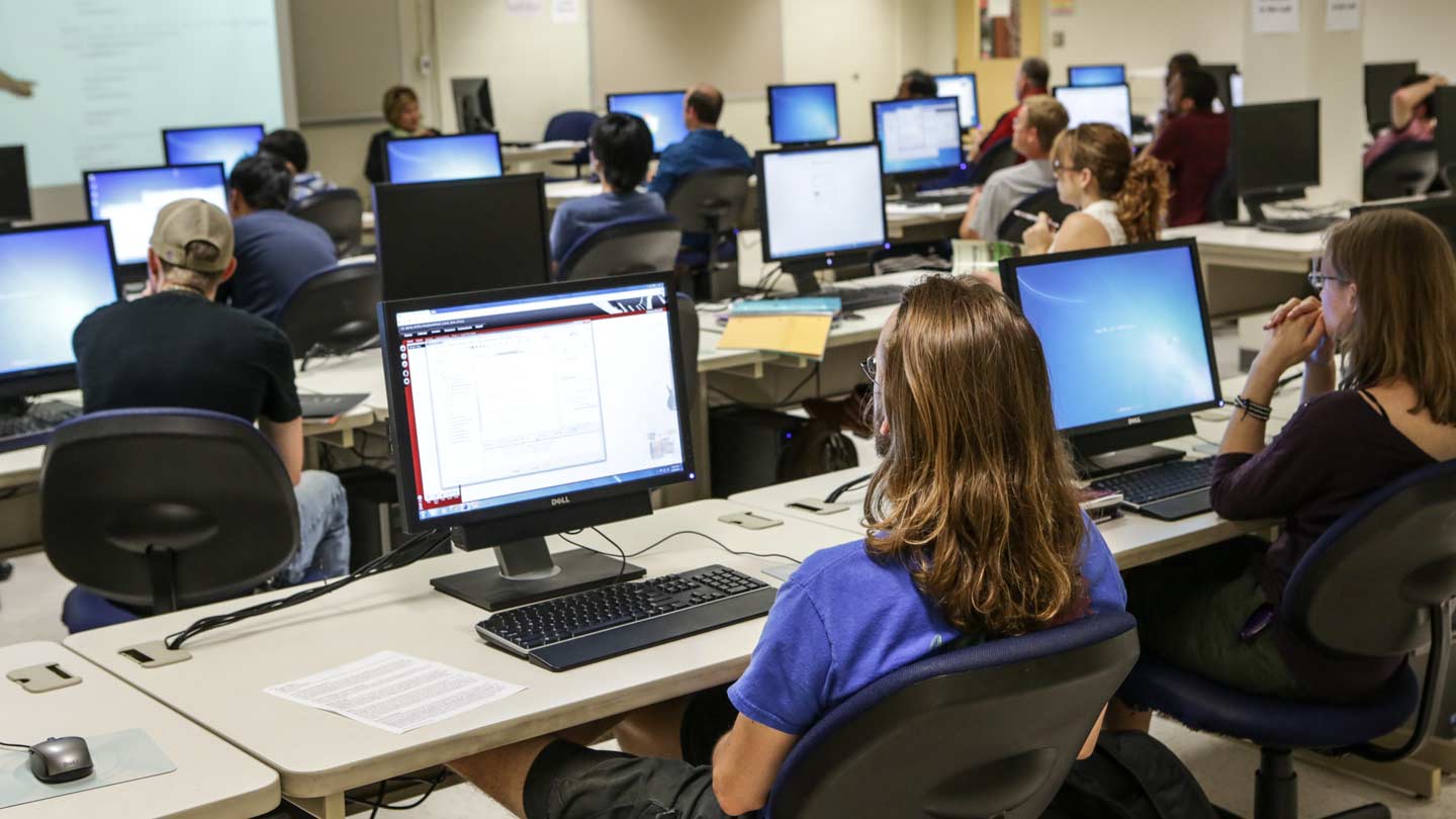 Students seated at computers in a classroom setting, focused on their screens during a lecture or instructional session.