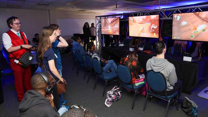 A large group of students gathers in a dimly lit room with festive neon lighting. They are watching multiple players play a game on three large TV screens.