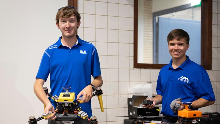 Two students in blue UAH polos pose indoors with a small yellow-and-black robot and orange equipment.