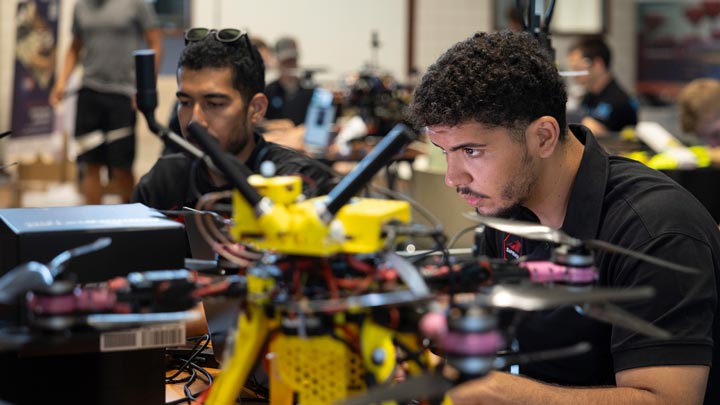 Two students assemble and test a yellow quadcopter drone at a workshop table.
