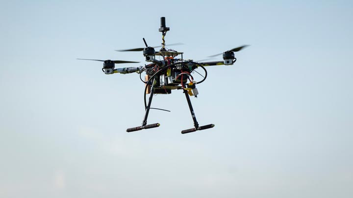 A homemade quadcopter drone with exposed wiring and landing skids hovers against a clear blue sky.