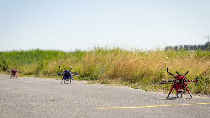 Three multirotor drones lined up on a rural paved road beside tall grass, with the nearest red drone in sharp focus.