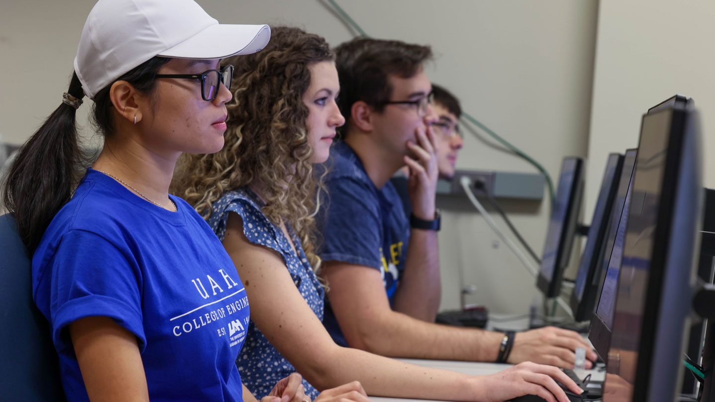 Three UAH students working on computers in a classroom, focused on their screens, wearing casual clothing.