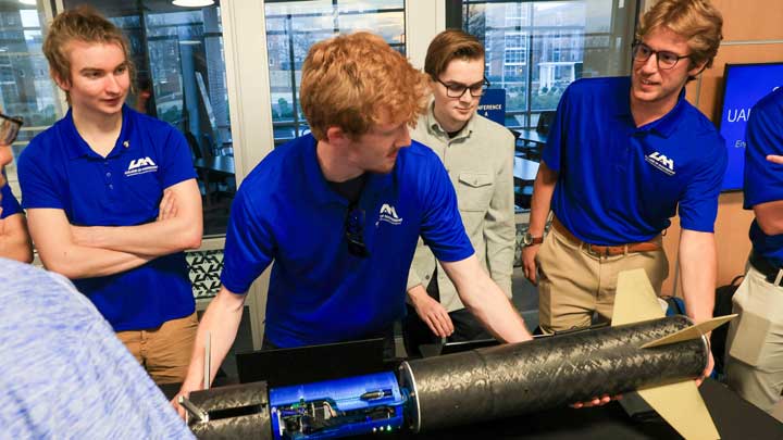 A group of UAH students in blue shirts examines a model rocket on a table, discussing its design and components.
