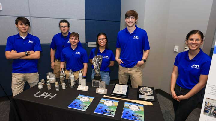A group of six individuals in blue shirts stands behind a table displaying various awards and jars, promoting their achievements.