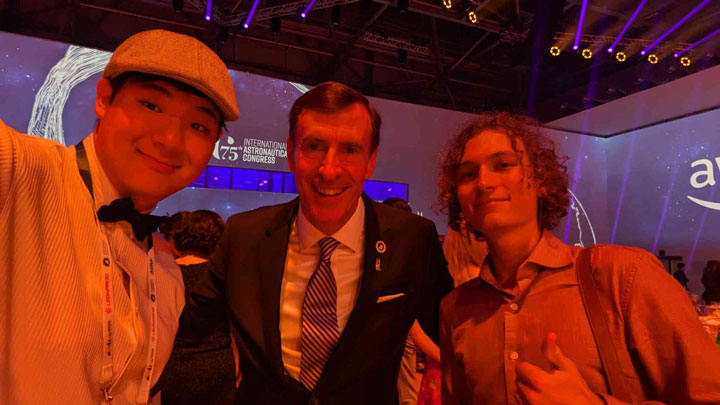 Three individuals at a lit event with a stage and the text International Astronautical Congress.