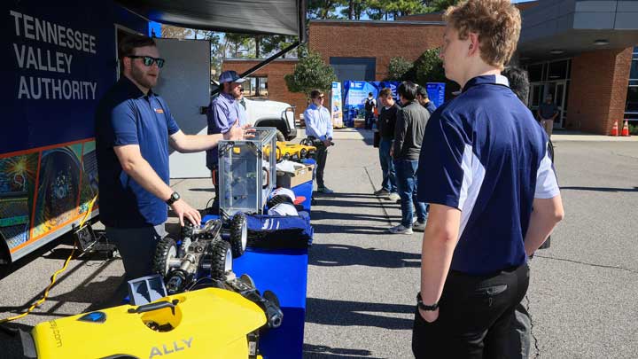 A group of people gathers around a display of robotic technology and equipment, discussing and demonstrating innovations outdoors.