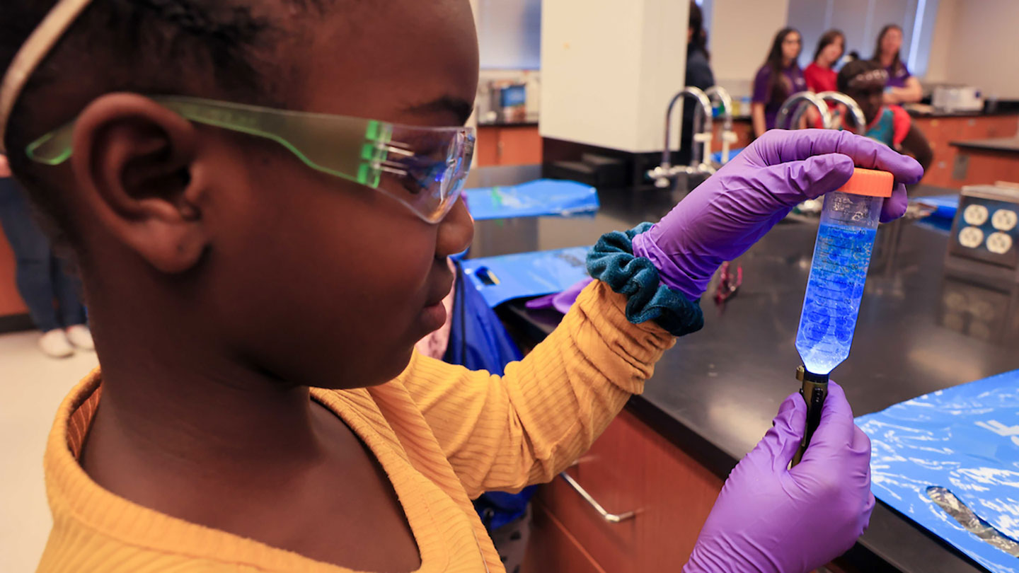 a young girl in safety goggles holds a beaker of blue liquid
