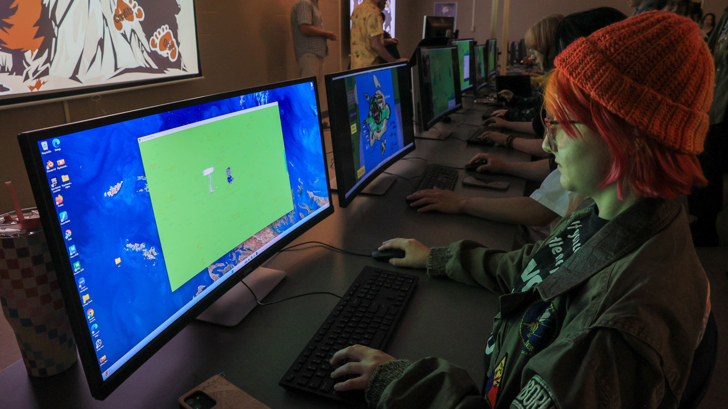 A group of individuals at computers in a dimly lit room, engaging with colorful video game screens, wearing casual attire.