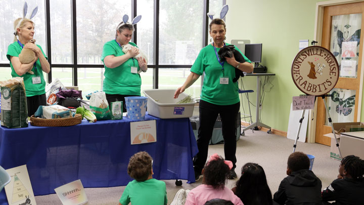 Three women in green shirts and bunny ear headbands hold rabbits, surrounded by pet supplies and a bright room with large windows.