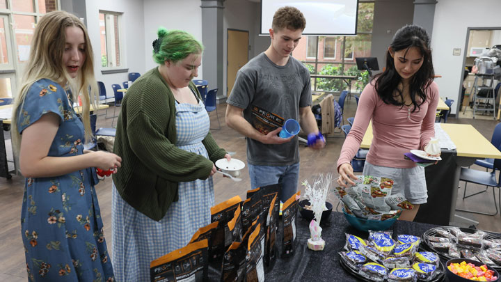 A group of four UAH students gathers around a table filled with snacks and treats in a brightly lit room.