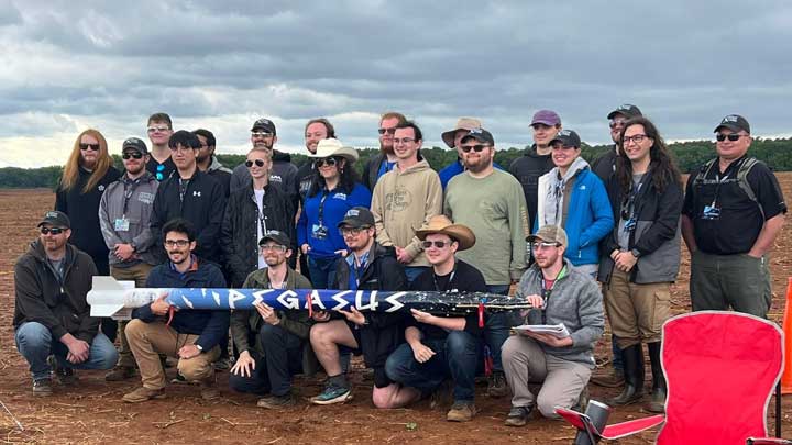 A group of diverse individuals poses together outdoors, holding a rocket labeled PEGASUS against a cloudy sky.