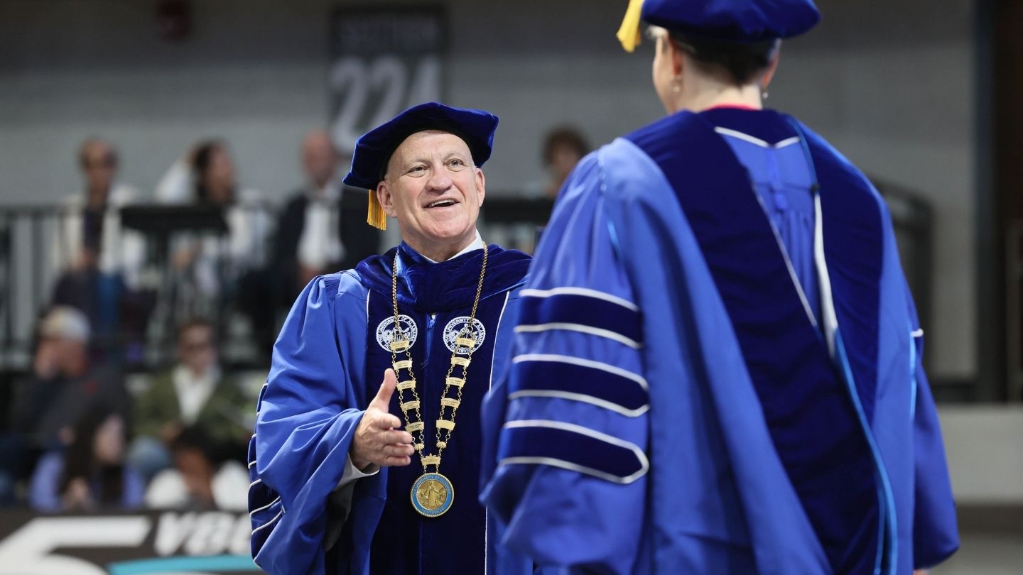 Dr. Karr, in academic robes, offers his hand to a student earning their doctorate.