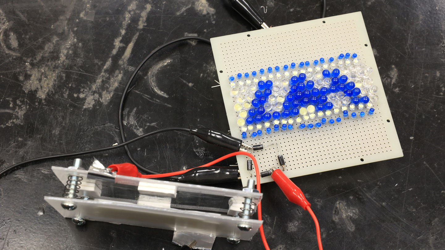 A circuit board displays UAH using colorful beads, with wires connected to a nearby component on a dark surface.