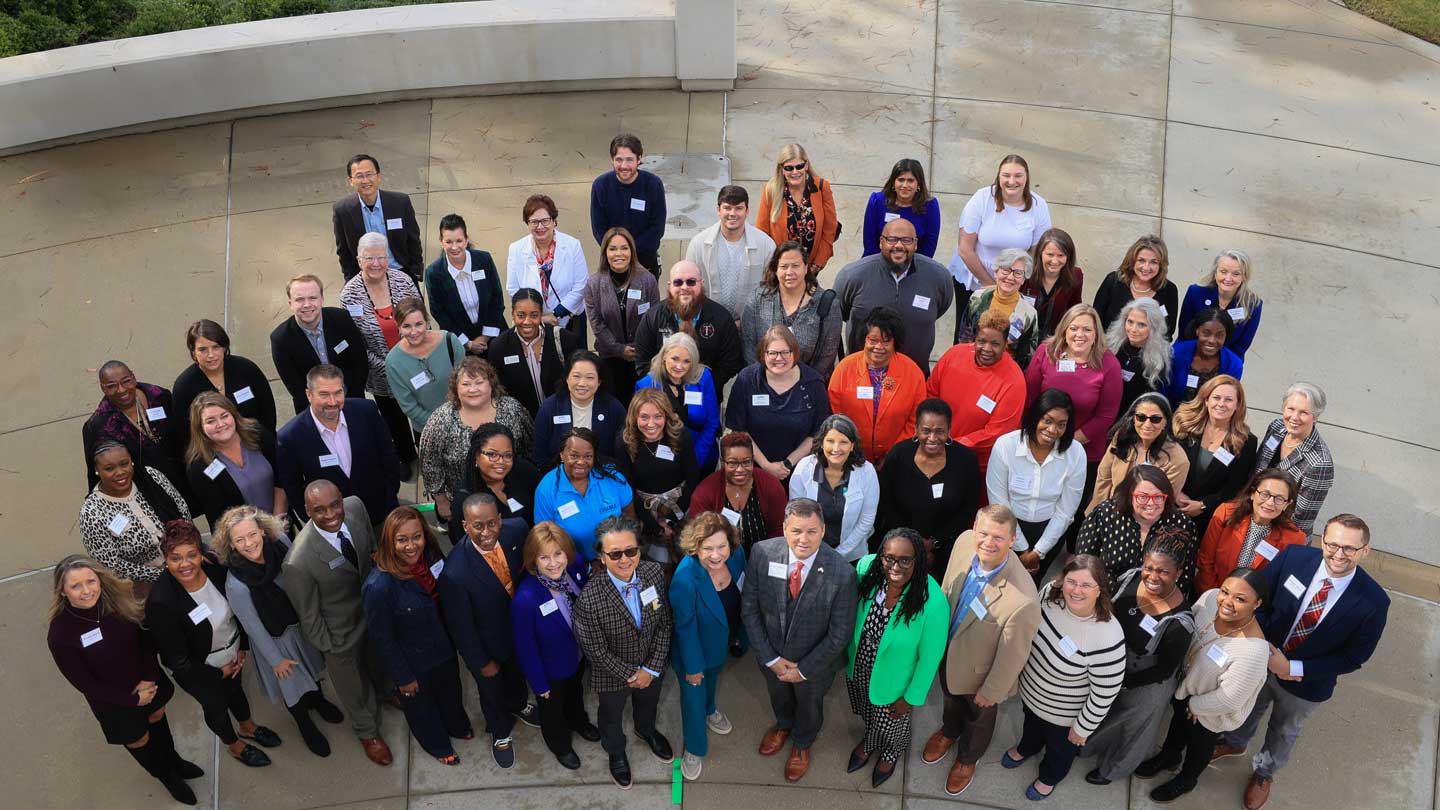 A large group of diverse individuals stands together outdoors, wearing name tags and a variety of professional attire.