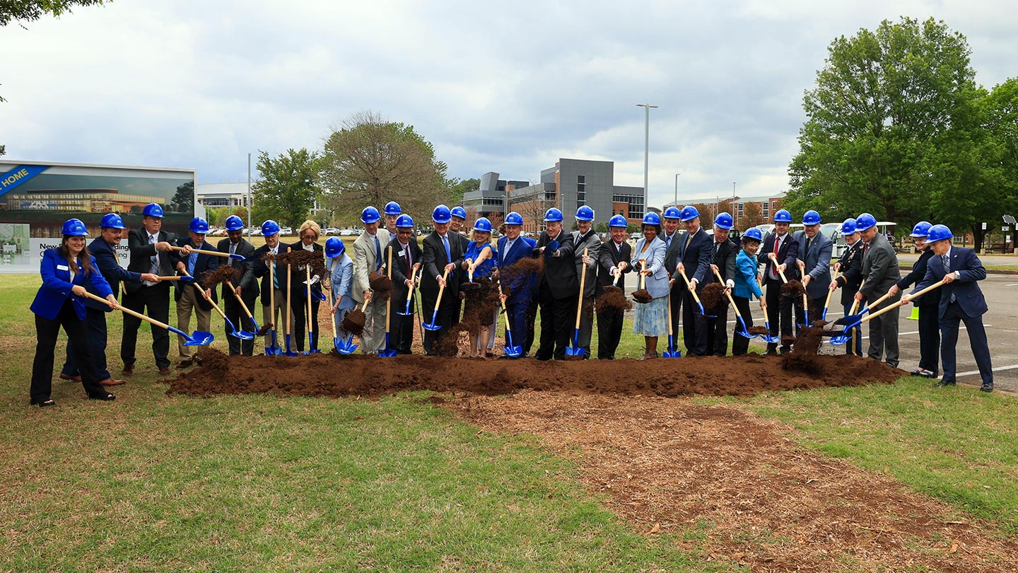 a large group of people in blue safety hats hold shovels near a pile of dirt