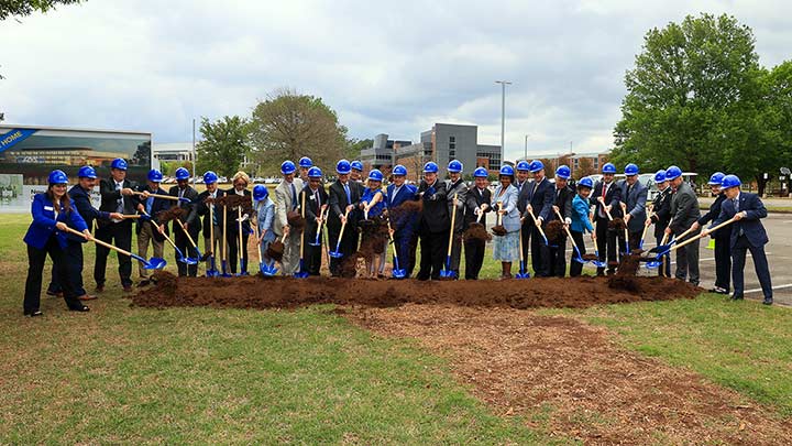 a large group of people in blue safety hats hold shovels near a pile of dirt ?>