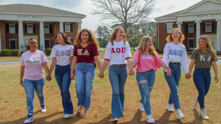 Seven women holding hands standing on grass in front of sorority building. ?>