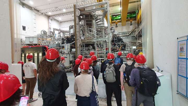 A group of visitors, wearing red hard hats, observes large industrial machinery in a spacious facility.