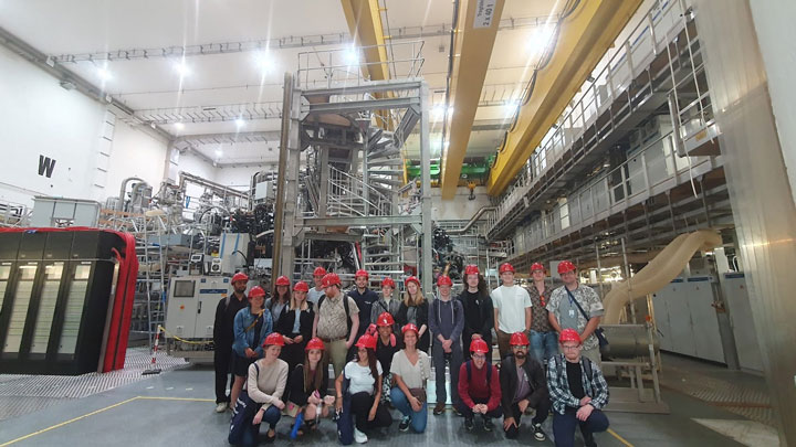 Group photo of visitors in hard hats in a large industrial facility, surrounded by machinery and equipment.