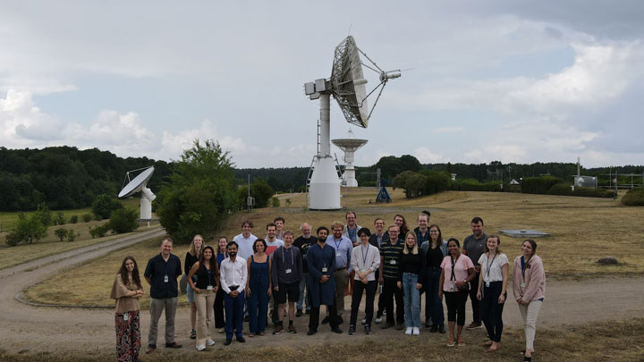A diverse group of 30 people stands in front of large satellite dishes against a cloudy sky, surrounded by green trees and a dirt path.