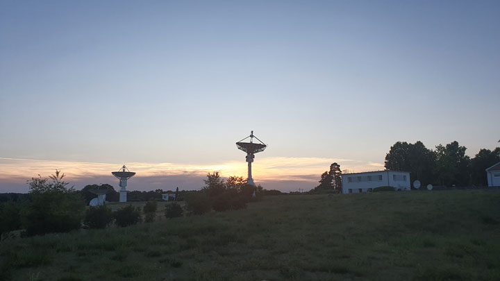 Silhouetted satellite dishes stand against a colorful sunset, with a grassy field and trees in the background.