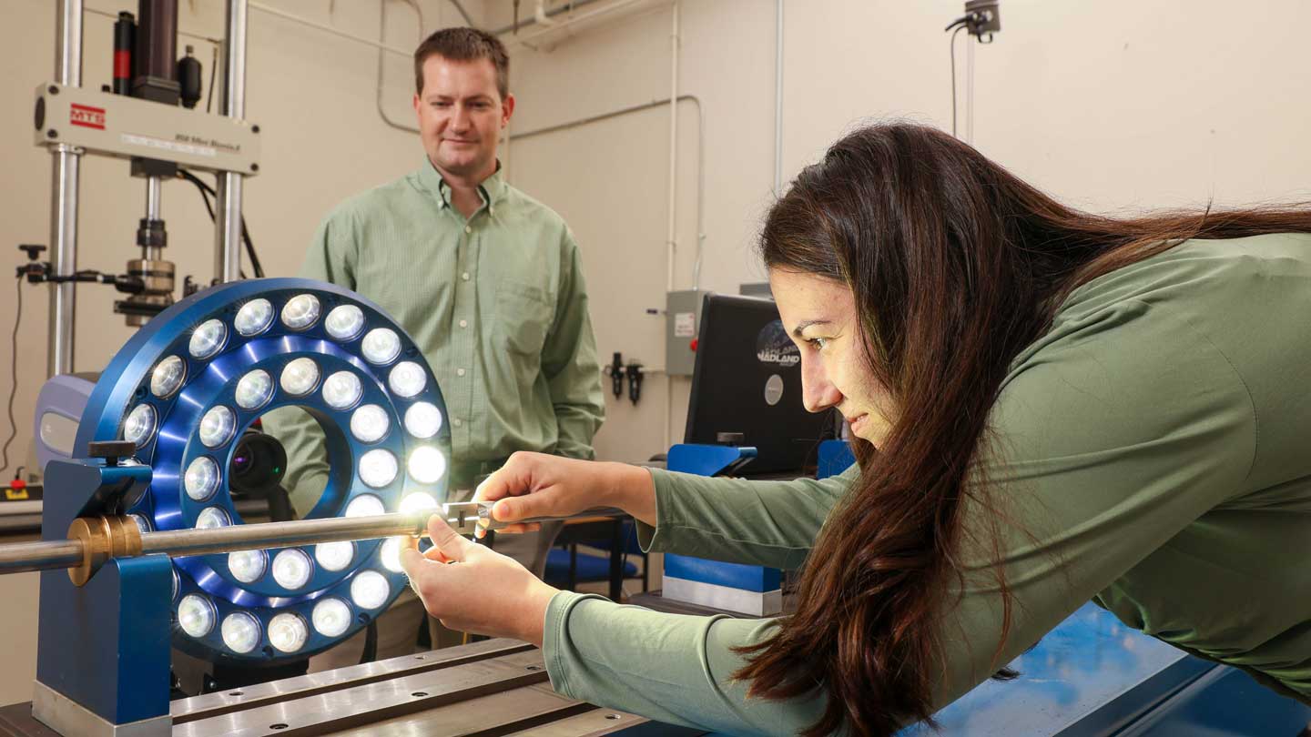A woman in a green shirt uses calipers on a metal rod, surrounded by equipment and a circular light ring in a laboratory setting.