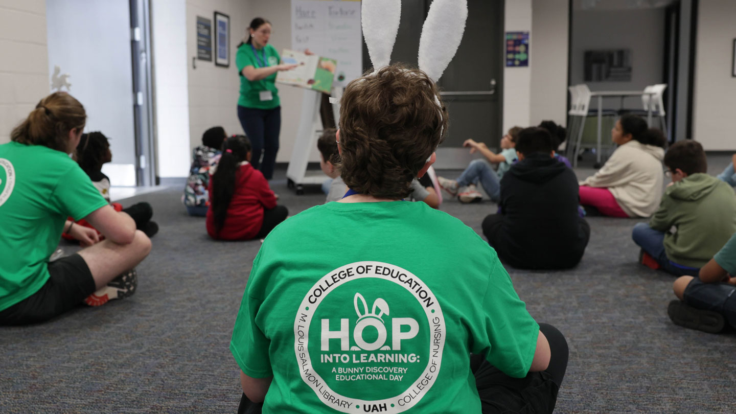 A group of children sits on the carpet, engaged in a learning activity led by a facilitator in a green shirt with bunny ears.