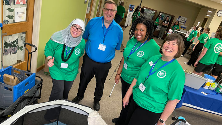 A group of four individuals in green shirts engages in discussion around a display at a community event, surrounded by informational booths.